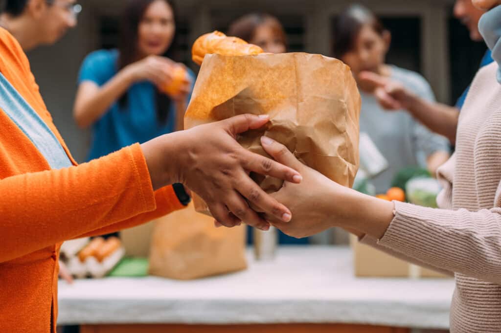 An unrecognizable woman hands a bag of donated food to someone during a community food drive. An Asian female volunteer and her colleagues distributing grocery food at community food bank